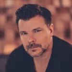 Portrait d’ATB, regard face caméra, dans une ambiance chaleureuse et intimiste, sur fond flou aux teintes brunes.