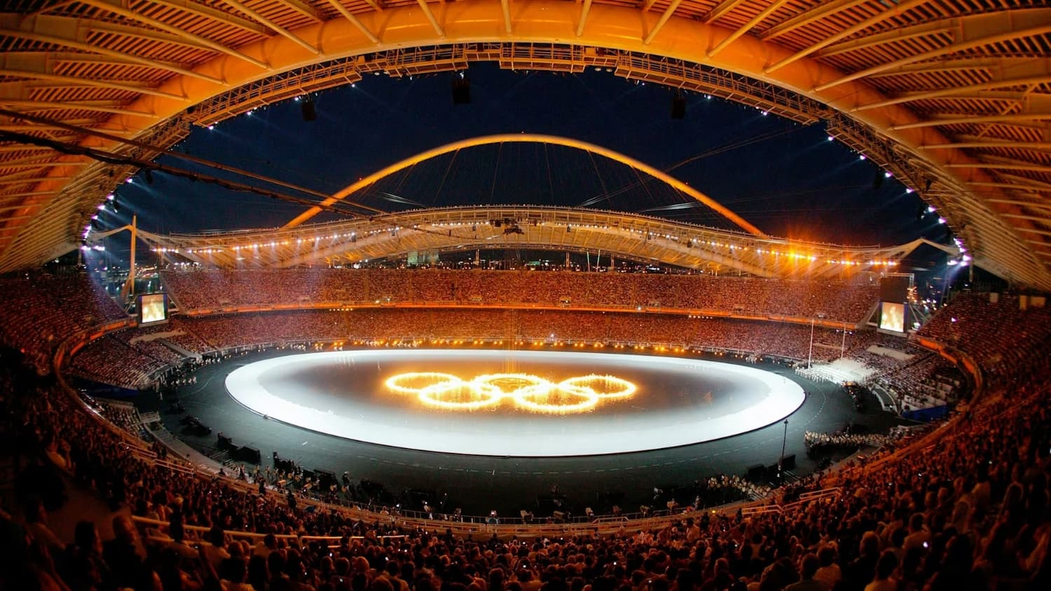 Stade olympique bondé lors des Jeux Olympiques d'Athènes 2004, avec les anneaux olympiques illuminés au centre de l'arène.