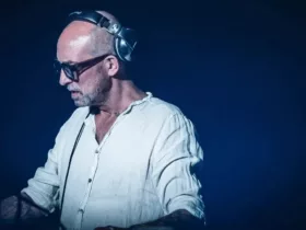 DJ Tomcraft se produisant sur scène, portant une chemise blanche, un casque et des lunettes, concentré sur son équipement.