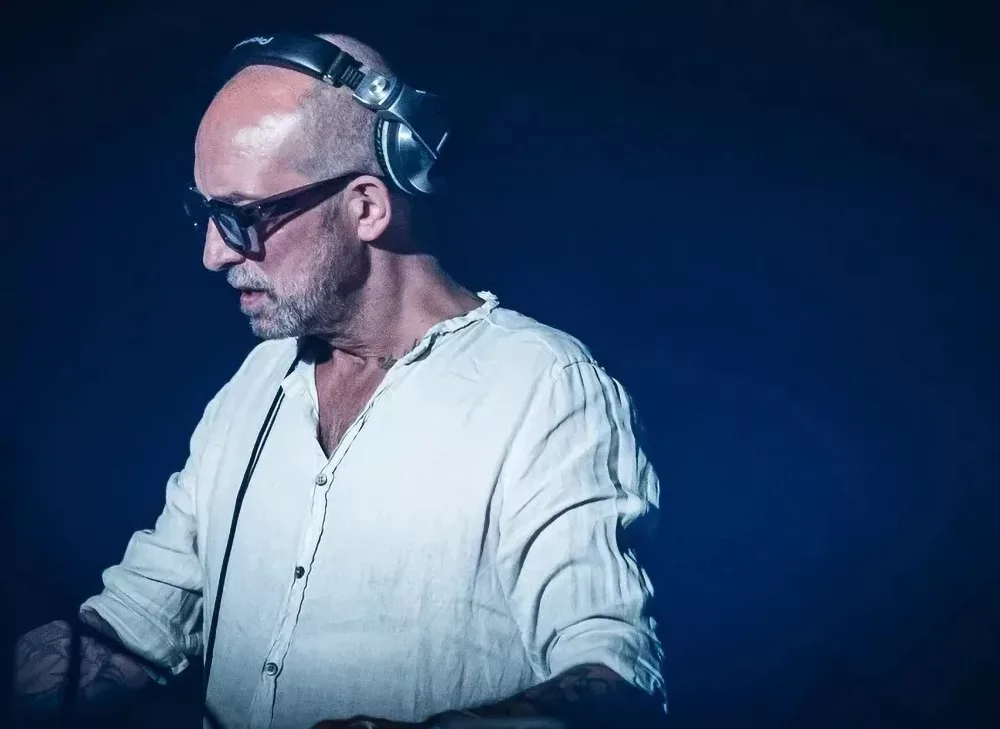 DJ Tomcraft se produisant sur scène, portant une chemise blanche, un casque et des lunettes, concentré sur son équipement.