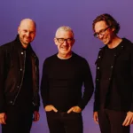 Above & Beyond posant ensemble sur fond violet, tous trois habillés en noir et souriants, lors de la promotion de l’album Bigger Than All Of Us.
