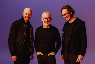 Above & Beyond posant ensemble sur fond violet, tous trois habillés en noir et souriants, lors de la promotion de l’album Bigger Than All Of Us.