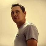 Tiësto en portrait, portant un t-shirt gris et regardant sur le côté, dans un éclairage chaud et minimaliste.