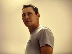 Tiësto en portrait, portant un t-shirt gris et regardant sur le côté, dans un éclairage chaud et minimaliste.