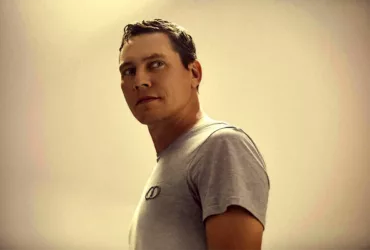 Tiësto en portrait, portant un t-shirt gris et regardant sur le côté, dans un éclairage chaud et minimaliste.