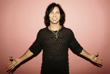 Photo presse de Skazi, souriant les bras ouverts, vêtu d’un haut noir texturé et d’un collier argenté.