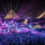 Festival électronique en plein air au pied des Pyramides de Gizeh, illuminé par des jeux de lasers et de lumière, avec une foule dense au cœur du désert égyptien. Il s'agit de "Future Sound of Egypt 500" par Aly & Fila.