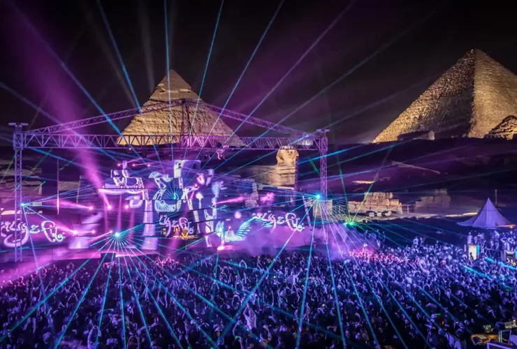 Festival électronique en plein air au pied des Pyramides de Gizeh, illuminé par des jeux de lasers et de lumière, avec une foule dense au cœur du désert égyptien. Il s'agit de "Future Sound of Egypt 500" par Aly & Fila.