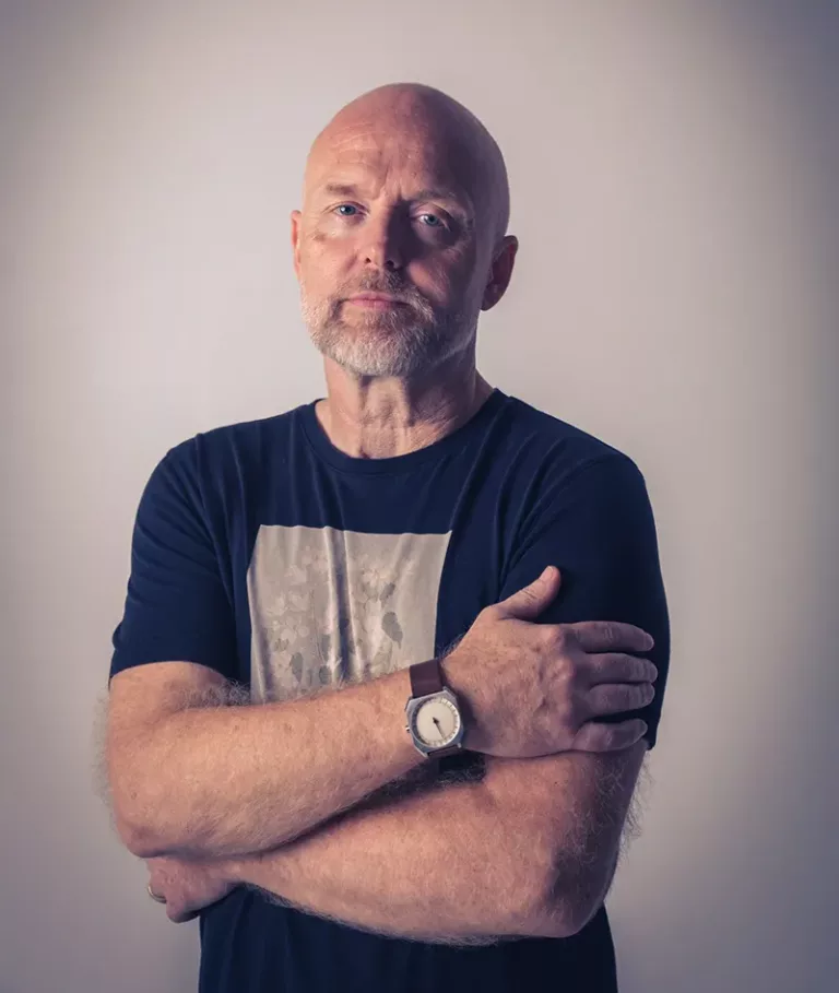 Portrait de Solarstone debout, les bras croisés, portant un t-shirt sombre et une montre au poignet, sur fond neutre éclairé.
