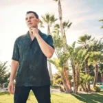 Portrait de Tiësto en extérieur, dans une ambiance ensoleillée et détendue, entouré de palmiers et de verdure.
