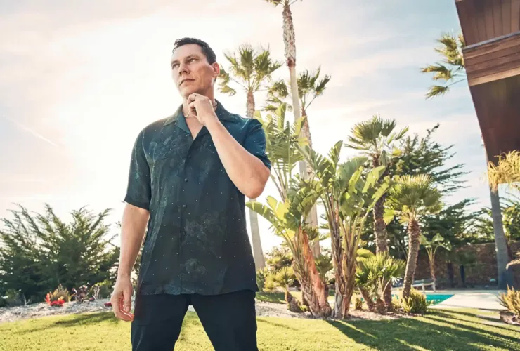 Portrait de Tiësto en extérieur, dans une ambiance ensoleillée et détendue, entouré de palmiers et de verdure.