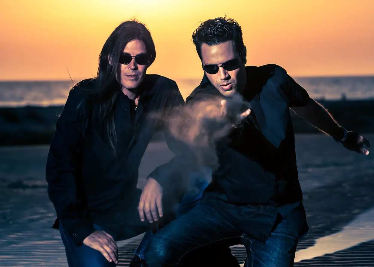 Le duo Astral Projection, figure emblématique de la Trance Goa, photographié sur une plage au coucher du soleil, dans une ambiance cinématique et mystique.