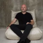 Solarstone assis sur un fauteuil beige, vêtu de noir, dans une pièce au mur brut en béton, pour un portrait minimaliste.