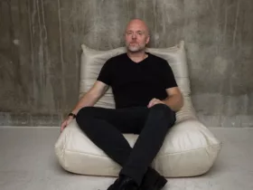 Solarstone assis sur un fauteuil beige, vêtu de noir, dans une pièce au mur brut en béton, pour un portrait minimaliste.