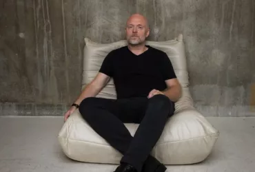 Solarstone assis sur un fauteuil beige, vêtu de noir, dans une pièce au mur brut en béton, pour un portrait minimaliste.