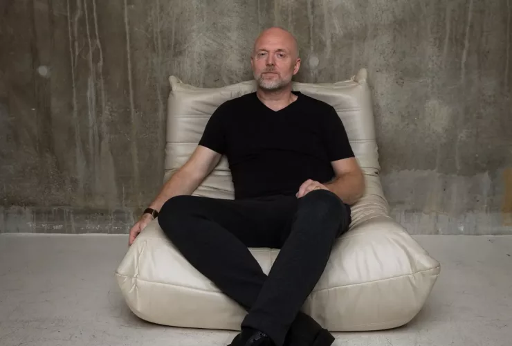 Solarstone assis sur un fauteuil beige, vêtu de noir, dans une pièce au mur brut en béton, pour un portrait minimaliste.