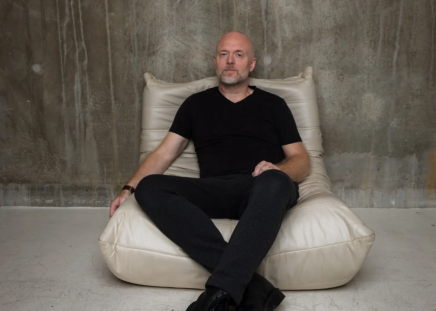 Solarstone assis sur un fauteuil beige, vêtu de noir, dans une pièce au mur brut en béton, pour un portrait minimaliste.