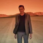 Tiësto debout dans le désert au coucher du soleil, baigné d’une lumière orange intense, vêtu d’un costume gris et d’un pull noir.
