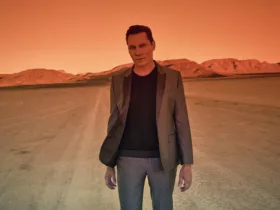 Tiësto debout dans le désert au coucher du soleil, baigné d’une lumière orange intense, vêtu d’un costume gris et d’un pull noir.