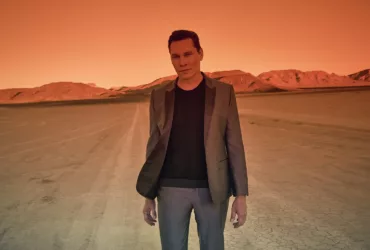 Tiësto debout dans le désert au coucher du soleil, baigné d’une lumière orange intense, vêtu d’un costume gris et d’un pull noir.