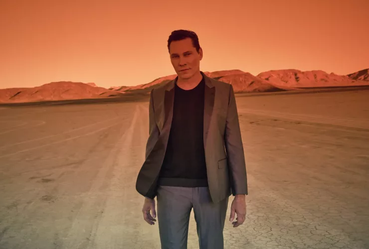 Tiësto debout dans le désert au coucher du soleil, baigné d’une lumière orange intense, vêtu d’un costume gris et d’un pull noir.