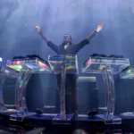 Tiësto sur scène, les bras levés, derrière un dispositif scénique futuriste transparent et lumineux, lors de son show Prismatic.