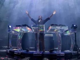 Tiësto sur scène, les bras levés, derrière un dispositif scénique futuriste transparent et lumineux, lors de son show Prismatic.