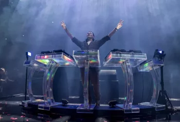 Tiësto sur scène, les bras levés, derrière un dispositif scénique futuriste transparent et lumineux, lors de son show Prismatic.