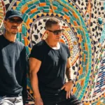 Le duo Cosmic Gate photographié devant une fresque en mosaïque colorée, dans une ambiance ensoleillée.