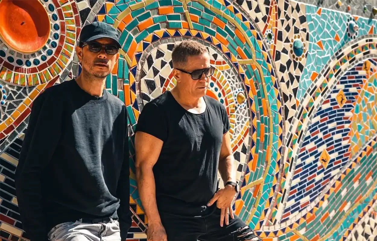 Le duo Cosmic Gate photographié devant une fresque en mosaïque colorée, dans une ambiance ensoleillée.