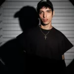 Mandragora en portrait devant un fond de type mugshot avec repères de taille, sous un éclairage contrasté créant une ombre marquée.