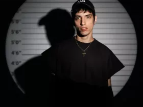 Mandragora en portrait devant un fond de type mugshot avec repères de taille, sous un éclairage contrasté créant une ombre marquée.