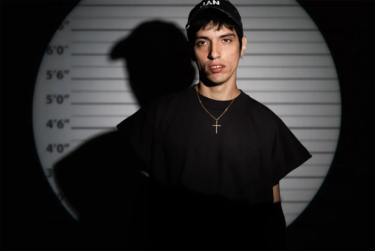 Mandragora en portrait devant un fond de type mugshot avec repères de taille, sous un éclairage contrasté créant une ombre marquée.