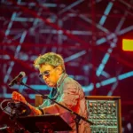 Jean-Michel Jarre en concert, manipulant ses instruments électroniques sur scène, sous un éclairage rouge et un décor lumineux.
