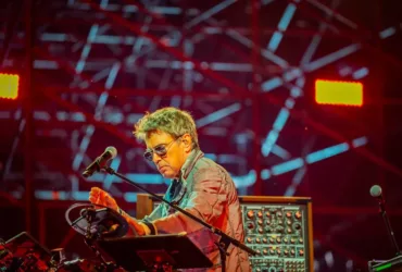 Jean-Michel Jarre en concert, manipulant ses instruments électroniques sur scène, sous un éclairage rouge et un décor lumineux.