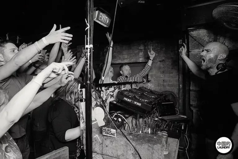 John 00 Fleming en DJ set dans un club bondé, interagissant avec le public levant les bras, en noir et blanc.
