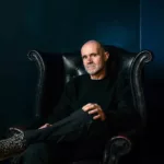 John 00 Fleming assis dans un fauteuil en cuir noir, les jambes croisées, dans une ambiance sombre aux tons bleu foncé.