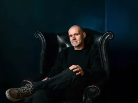 John 00 Fleming assis dans un fauteuil en cuir noir, les jambes croisées, dans une ambiance sombre aux tons bleu foncé.