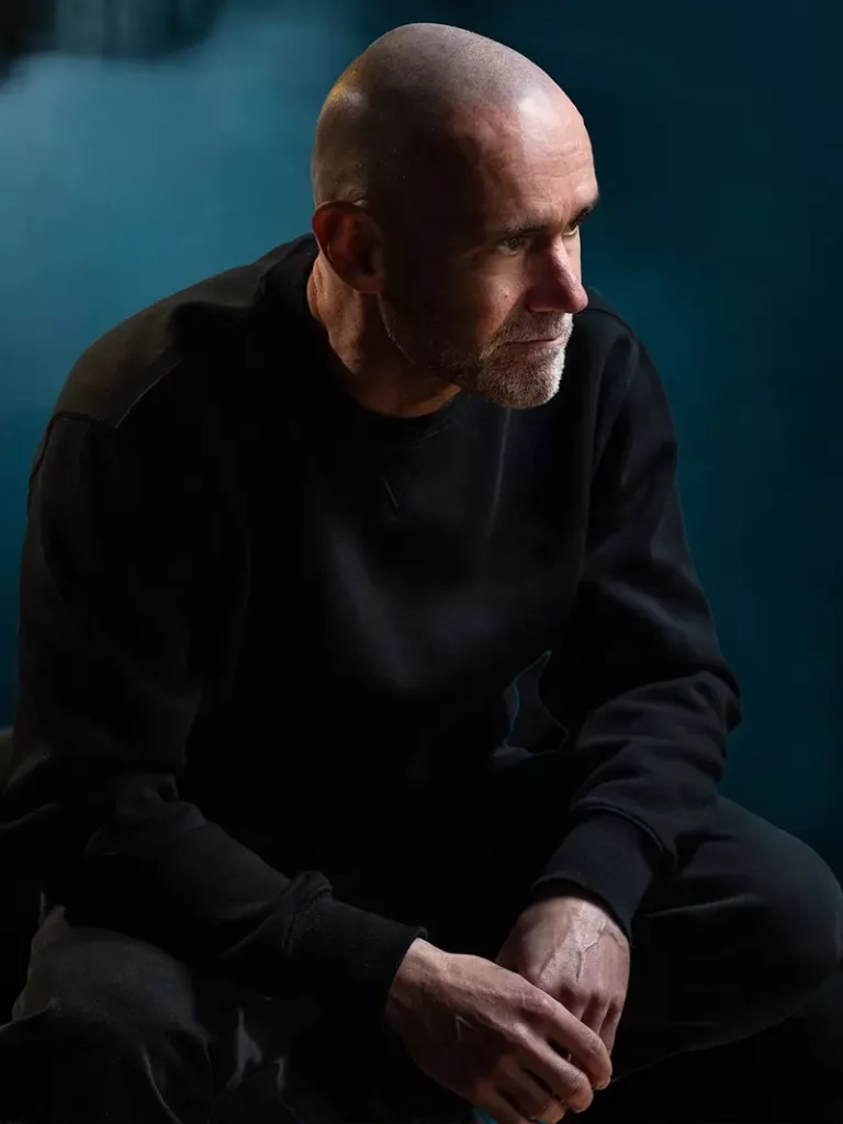 John 00 Fleming assis, regard tourné sur le côté, dans une ambiance sombre aux tons bleu foncé.