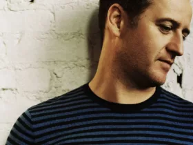 Portrait de Sasha, figure de la Progressive House & Trance, regard baissé, devant un mur en briques blanches.