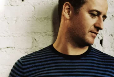 Portrait de Sasha, figure de la Progressive House & Trance, regard baissé, devant un mur en briques blanches.