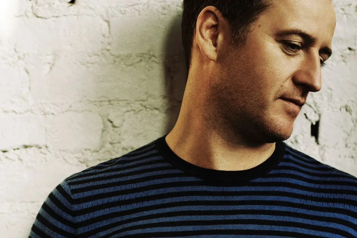 Portrait de Sasha, figure de la Progressive House & Trance, regard baissé, devant un mur en briques blanches.