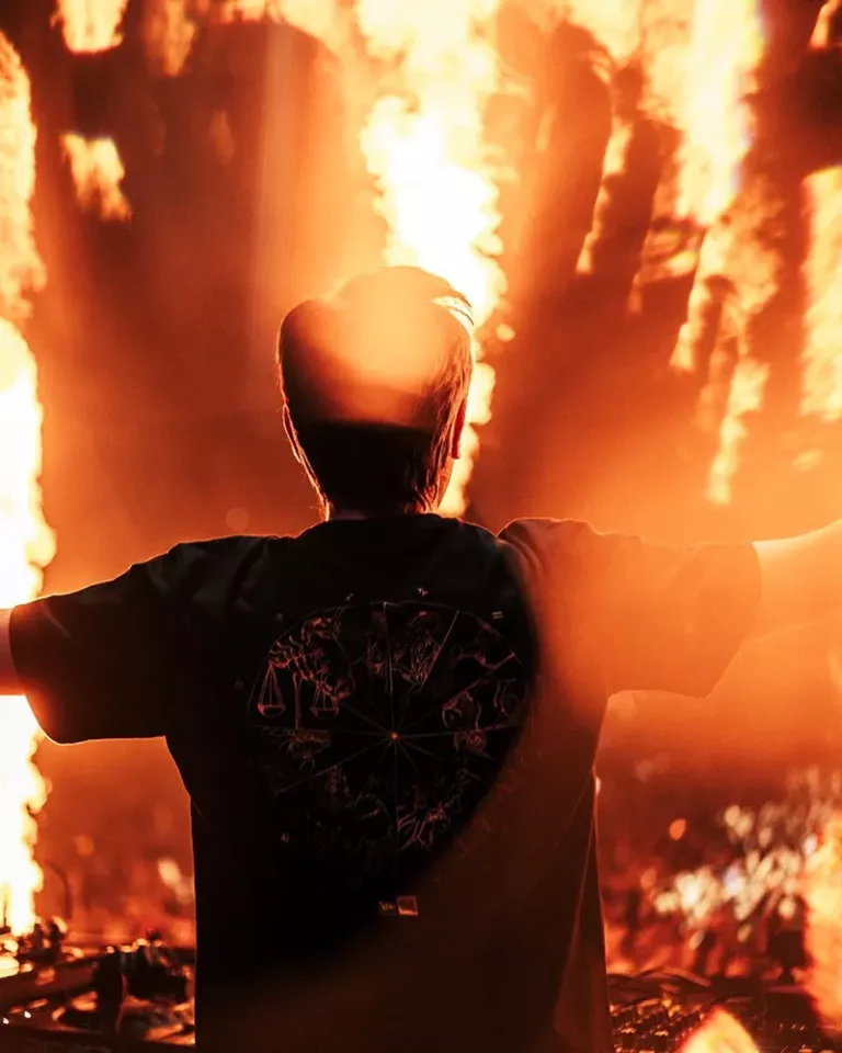 Andrew Rayel de dos sur scène, les bras ouverts face à la foule, entouré de jets de flammes et d’une lumière orange intense lors de son set à Dreamstate Europe 2025 en Pologne.
