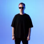 Andrew Rayel debout face caméra, portant des lunettes de soleil et un t-shirt noir, sur fond dégradé bleu avec un éclairage aux teintes froides.