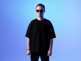 Andrew Rayel debout face caméra, portant des lunettes de soleil et un t-shirt noir, sur fond dégradé bleu avec un éclairage aux teintes froides.