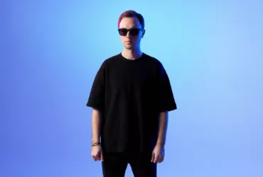 Andrew Rayel debout face caméra, portant des lunettes de soleil et un t-shirt noir, sur fond dégradé bleu avec un éclairage aux teintes froides.