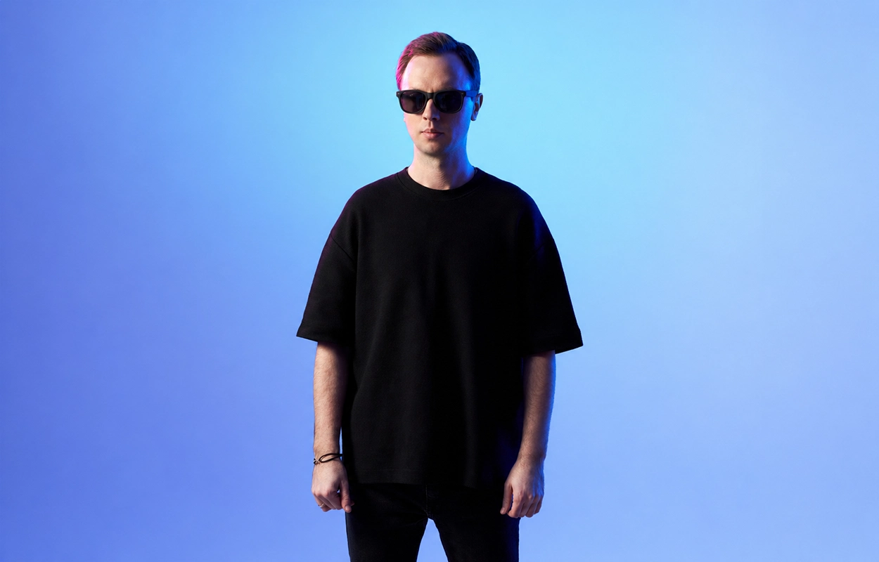 Andrew Rayel debout face caméra, portant des lunettes de soleil et un t-shirt noir, sur fond dégradé bleu avec un éclairage aux teintes froides.