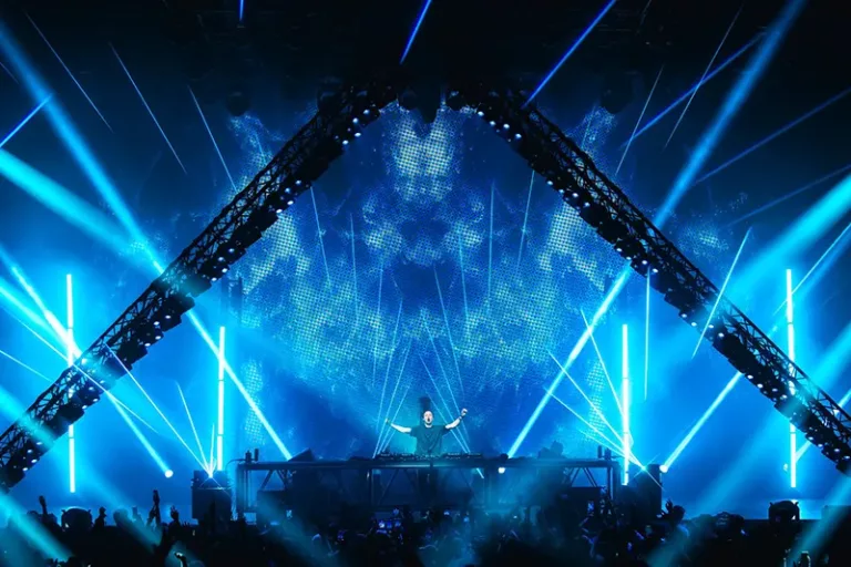 Andrew Rayel sur scène à Kuala Lumpur, les bras levés derrière ses platines, entouré de lasers bleus et d’une scénographie imposante en forme de triangle.