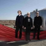 Le trio Above & Beyond pose devant deux caravanes argentées dans un paysage désertique, vêtu de noir et entouré d'un long voile rouge qui traverse le premier plan de la scène.