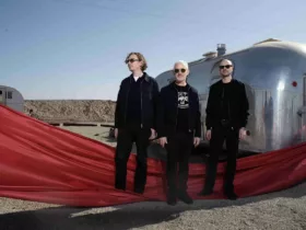 Le trio Above & Beyond pose devant deux caravanes argentées dans un paysage désertique, vêtu de noir et entouré d'un long voile rouge qui traverse le premier plan de la scène.